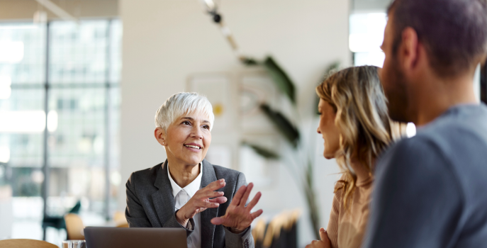 Professional female with short white hair sitting and talking to a young couple with their backs’ turned